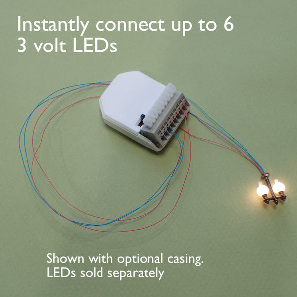 A white electronic module with attached red and blue wires, shown on a light green background with text reading 'Instantly connect up to 6 3 volt LEDs' and 'Shown with optional casing. LEDs sold separately.